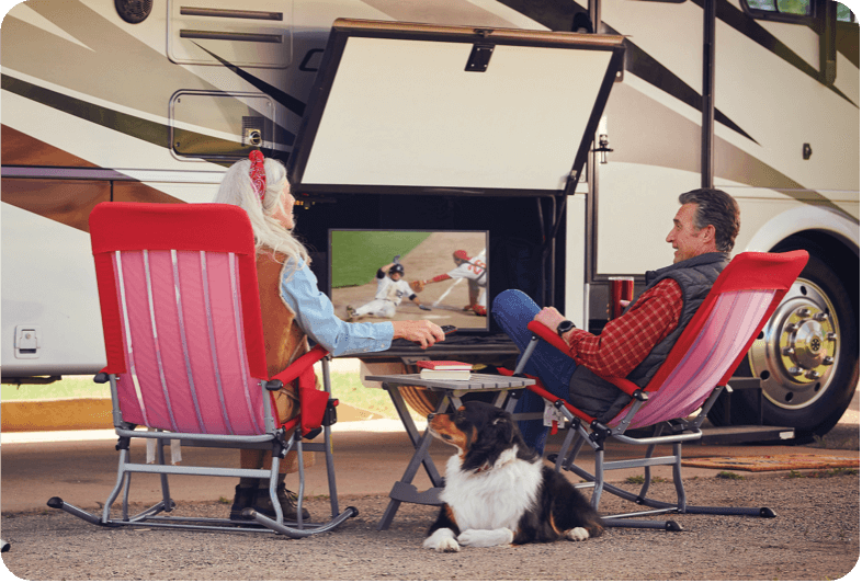 Couple sitting on chairs outside the RV, watching television with their dog between them.